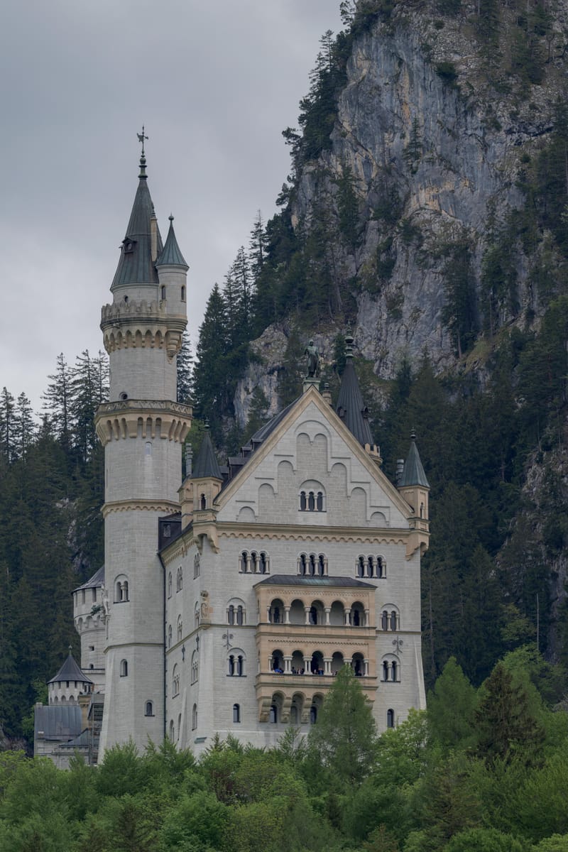 Neuschwanstein Castle rising from the forest below a grey cliff.