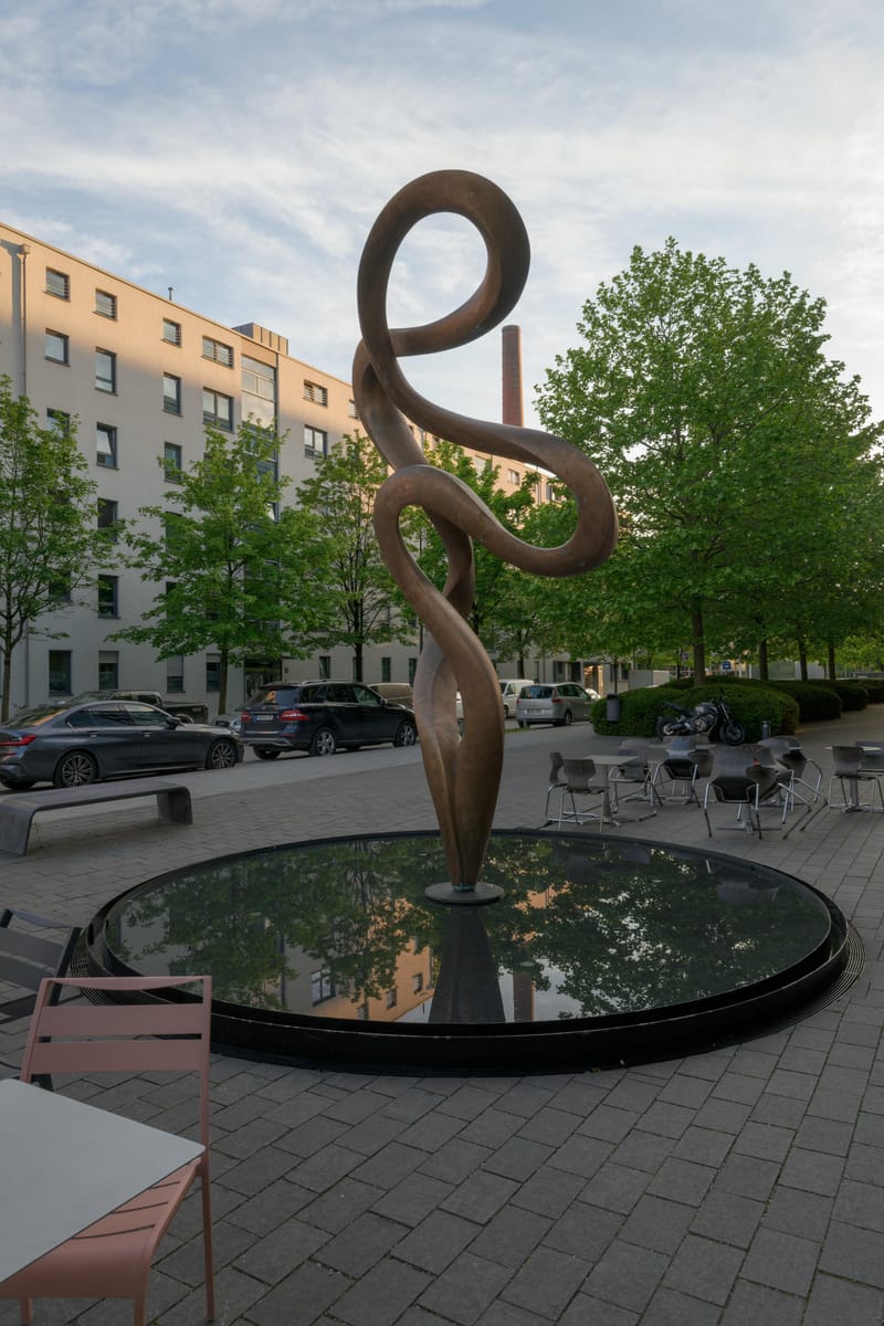 Looping bronze sculpture standing in a dark round fountain on a plaza.