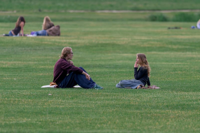 Two young people sitting face to face on a wide lawn, talking, with sunbathers behind them.