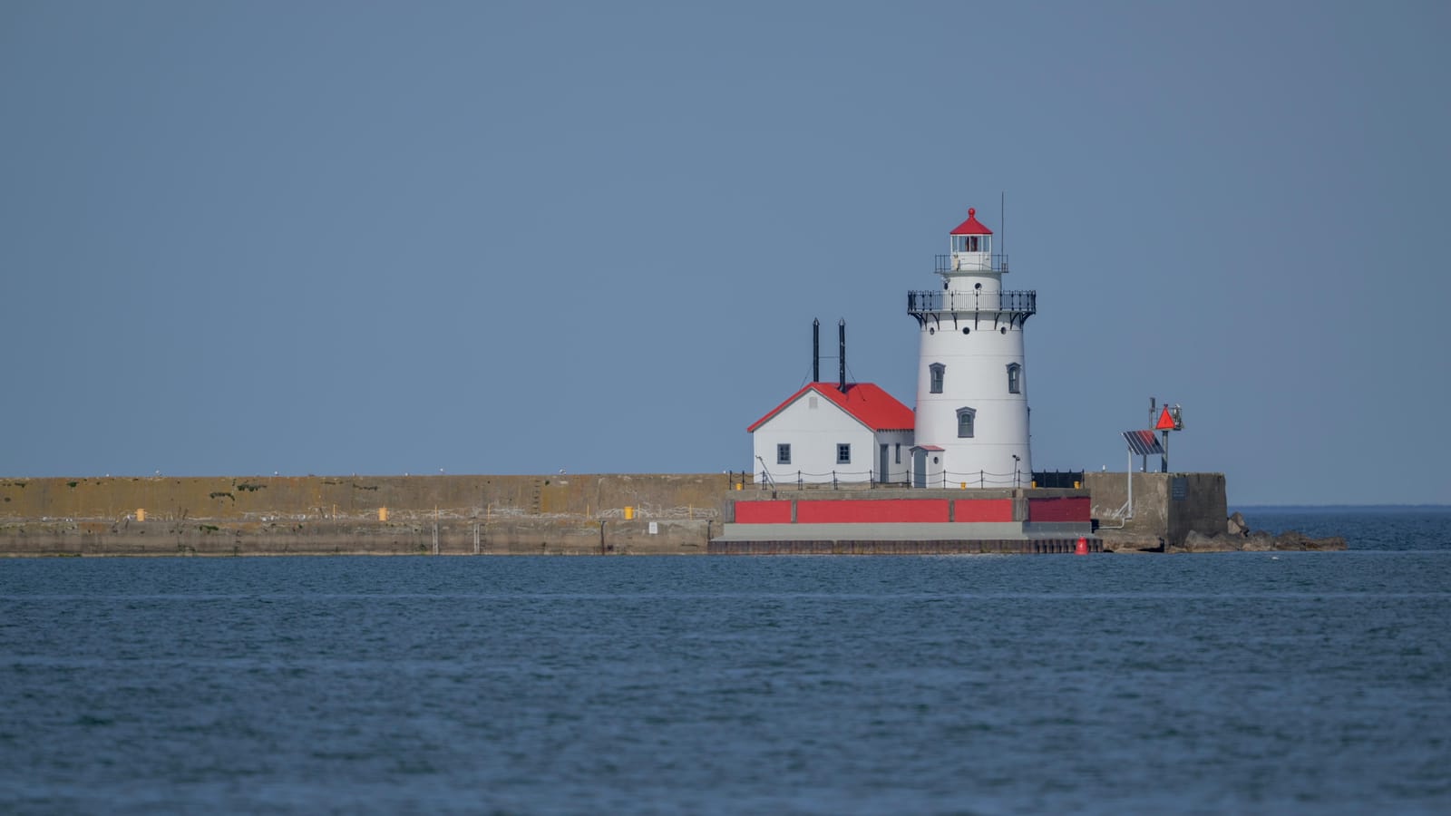 White lighthouse with a red roof at the end of a breakwater on calm blue water.