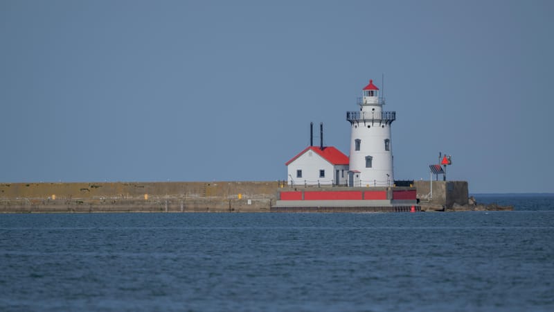 White lighthouse with a red roof at the end of a breakwater on calm blue water.