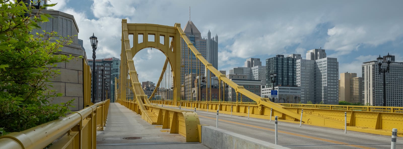 Yellow suspension bridge leading into downtown Pittsburgh under scattered clouds.