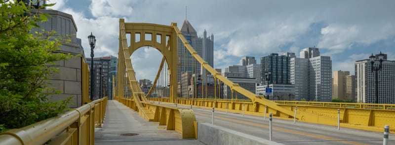 Yellow suspension bridge leading into downtown Pittsburgh under scattered clouds.